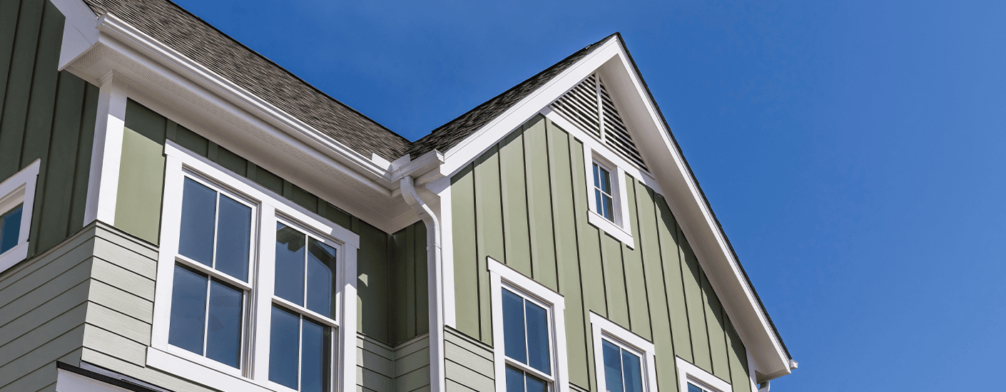 Close-up of the upper corner of a green board-and-batten home exterior with white trim, gutters, and windows against a clear blue sky.